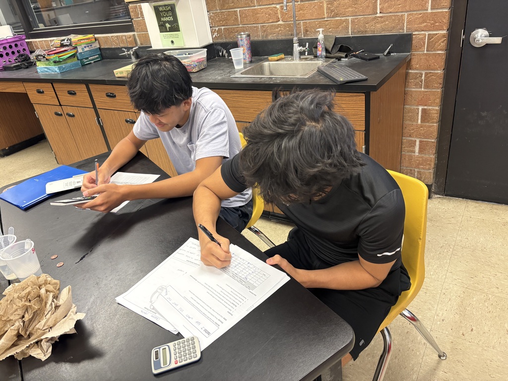 Two students seated at a table, one with a calculator and paper, the other writing, in a classroom.