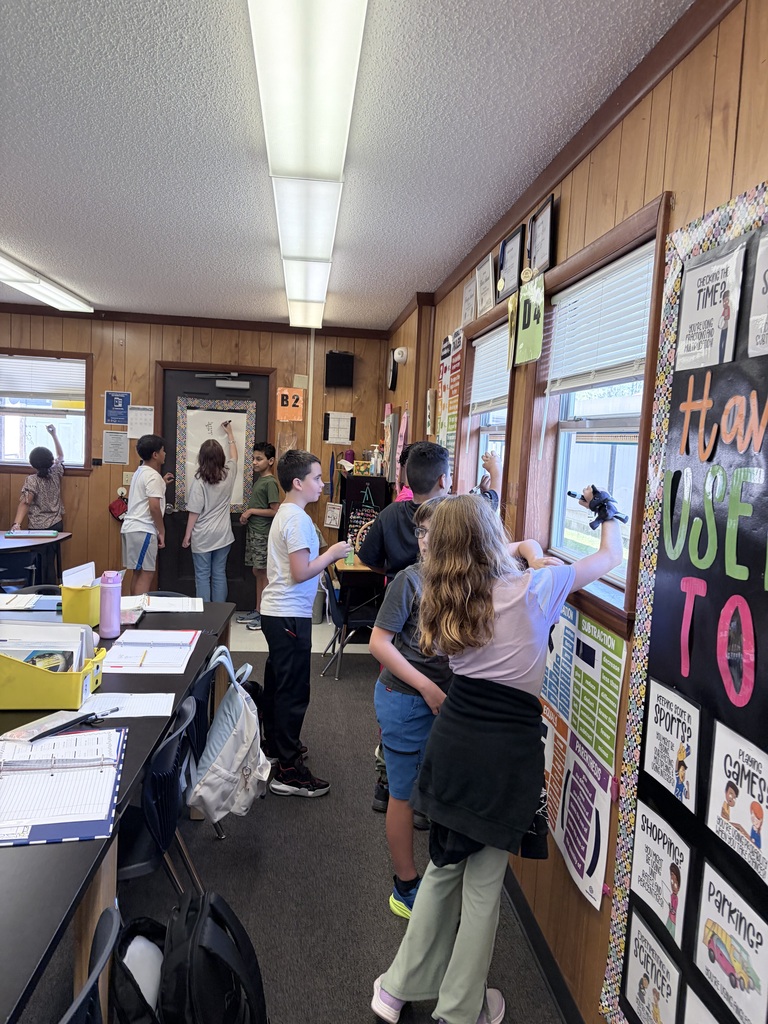 People are in a classroom; some are looking out the window and one is writing on a blackboard.