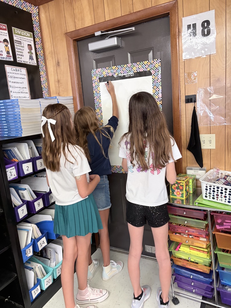 Three students write on a whiteboard in a classroom with wooden walls and storage shelves.