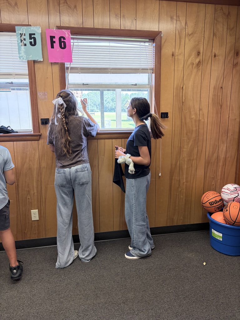 Two people stand by a window in a room with wooden walls. One wears a white cloth, and the other holds a towel.