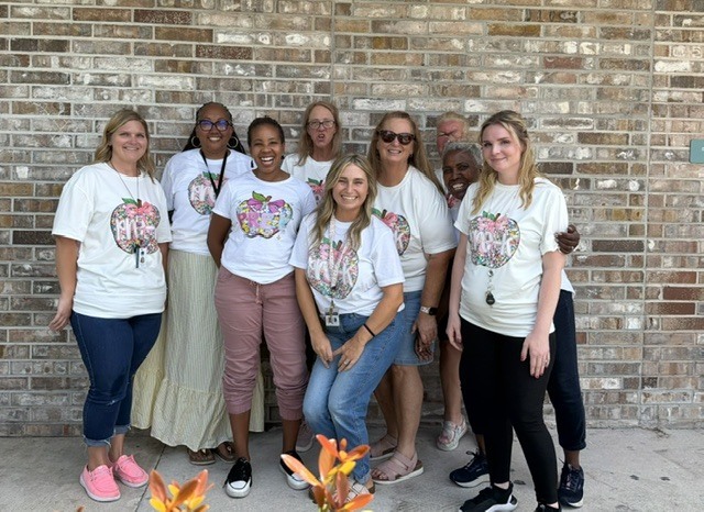 A group of adults in matching white T-shirts stands in front of a brick wall.