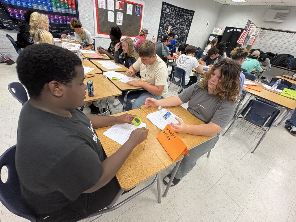 A classroom with students sitting at desks, some wearing bracelets. A teacher assists a student.