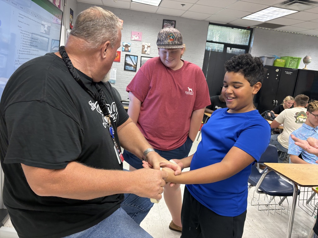 A man and a young boy shake hands in a classroom. Other students are seated at tables behind them.