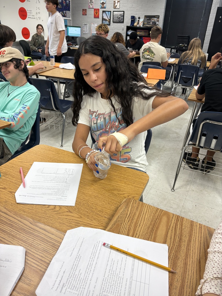 A girl in a white t-shirt works at a table with a glass of liquid and papers.