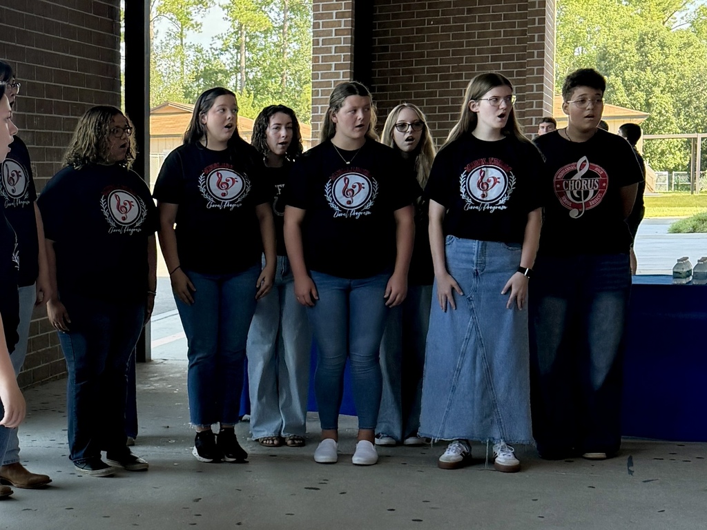 A group of young people in matching black t-shirts stand in front of a brick wall. Some wear denim pants; one wears a long skirt.