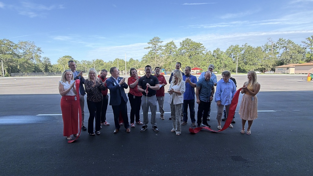 Group of people in formal wear standing on a road, holding a red ribbon for a ribbon-cutting event.