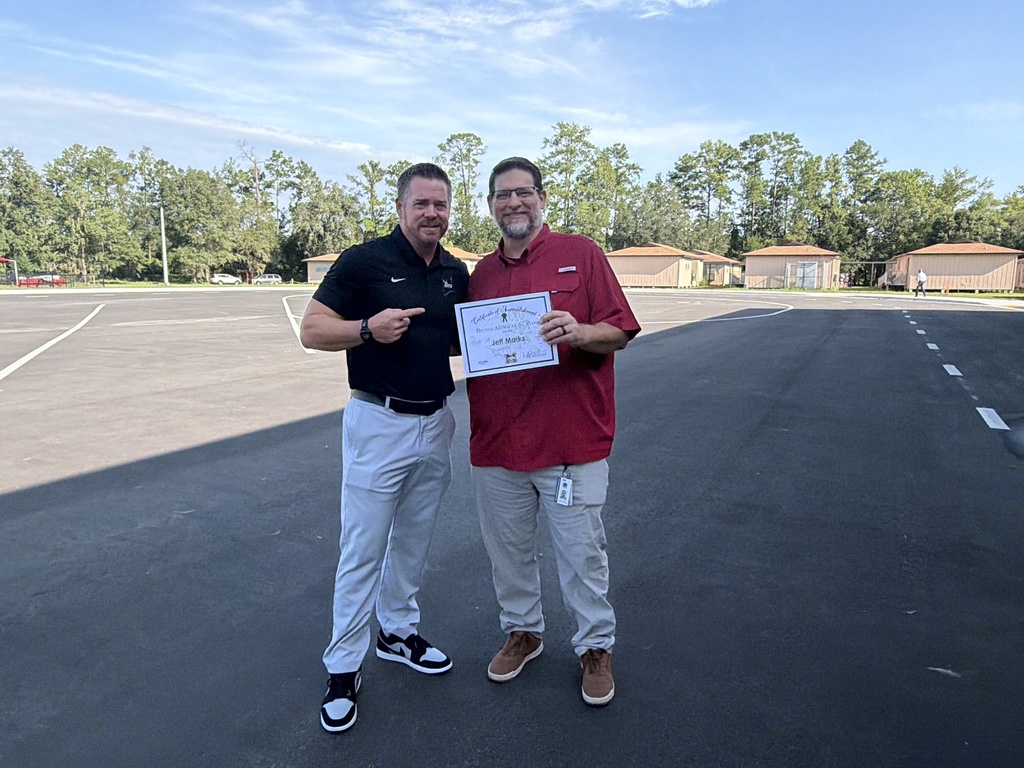 Two men stand in a parking lot. One holds a certificate. The other points. Trees and houses in the background.