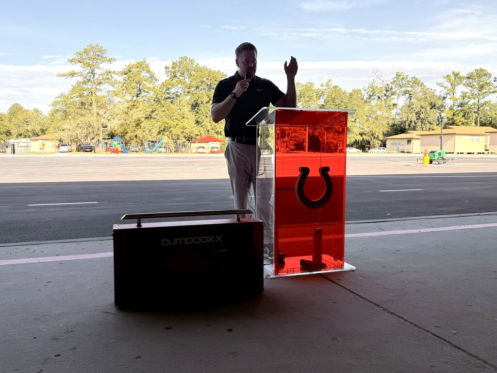 Person with a microphone stands at a podium with a red horseshoe design on a paved area.