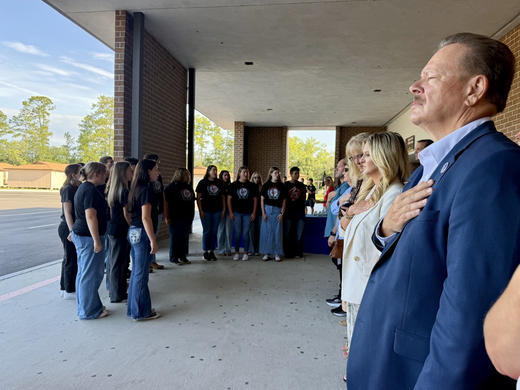 A group of people stands outside a building; a man in a blue suit has his hand over his heart.