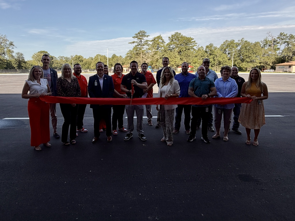 A group of people stand behind a red ribbon on an asphalt road with trees and a building in the distance.