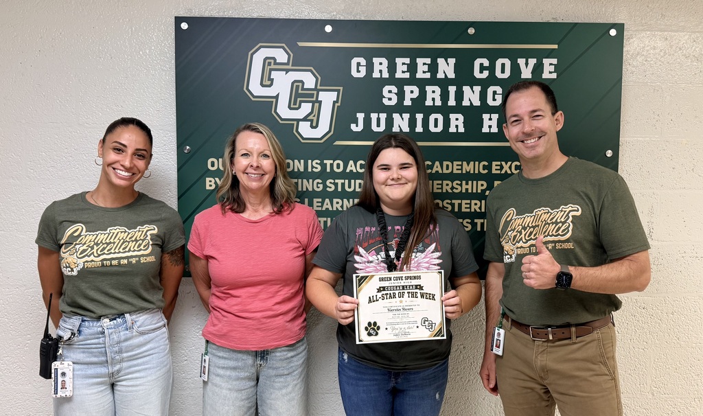 Four adults stand together in front of a green sign. The two women hold a certificate. The sign reads Green Cove Spring Junior High.