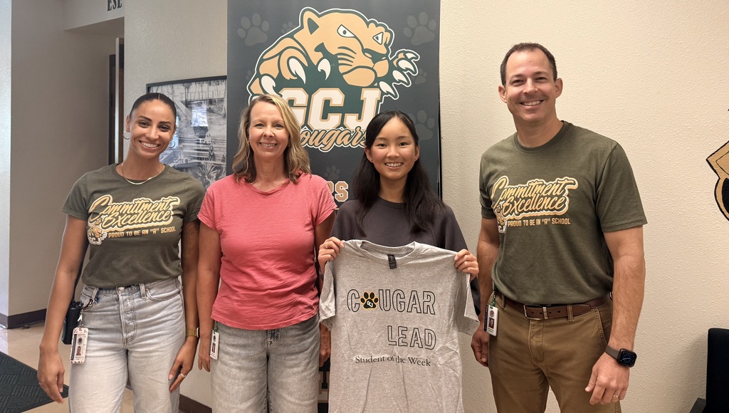 Four people stand with a shirt between two women. They are in front of a wall with a banner.