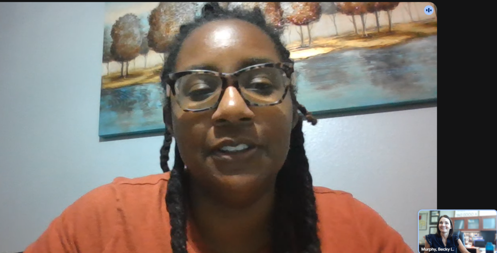 A woman with braided hair and glasses smiles, against a background featuring a landscape painting.