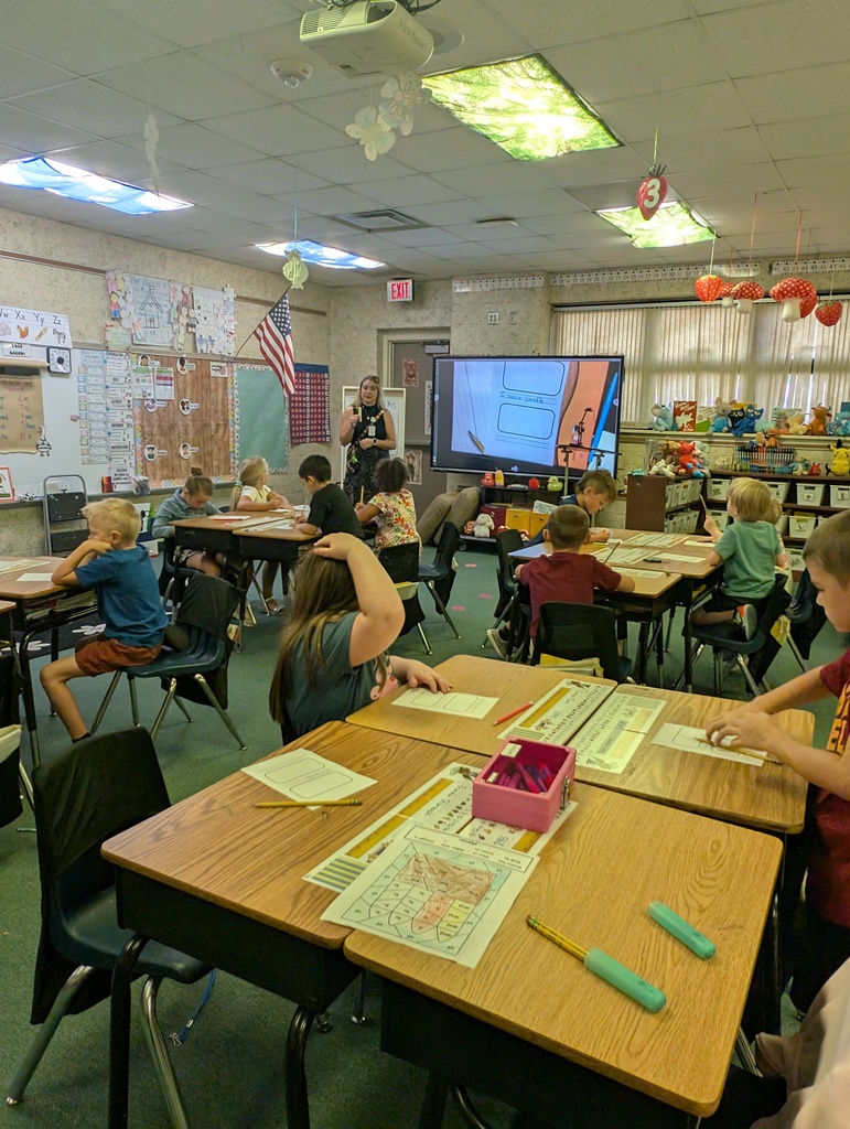Classroom with children seated at desks, a teacher standing near a projector screen. Desks and chairs are arranged in rows.