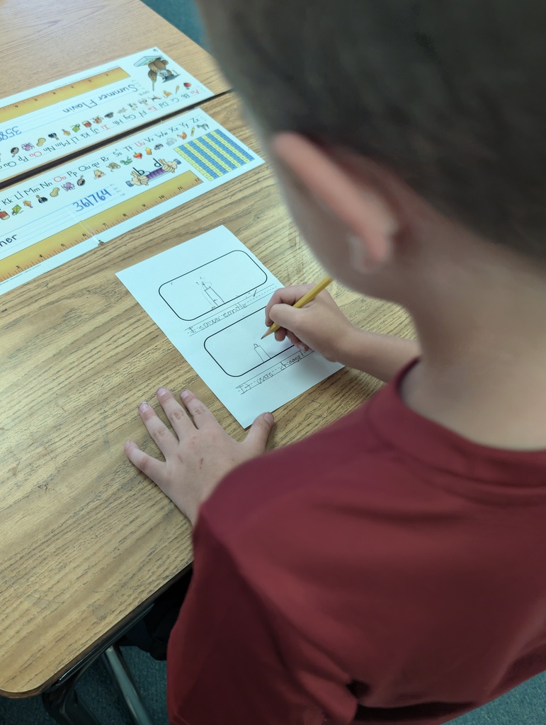 A boy in a red shirt sits at a desk, writing on a paper with a pencil.