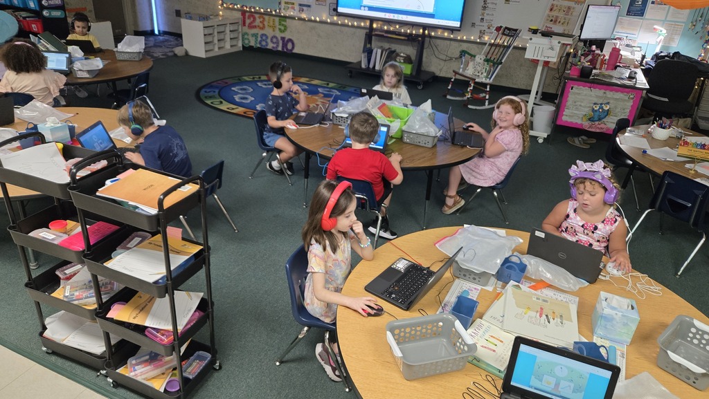 A classroom with students seated at round tables, using laptops and headphones, and a teacher monitor on the wall.