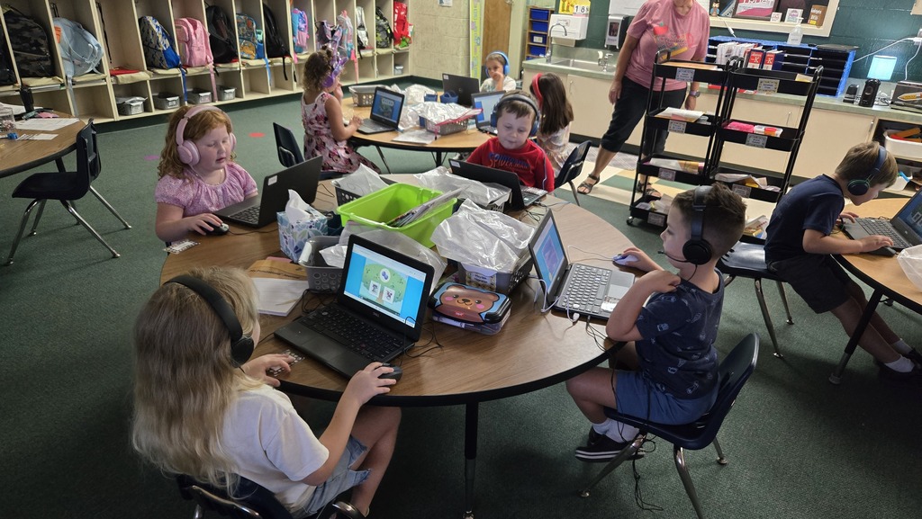 Children in a classroom, wearing headphones, using laptops at tables. One adult standing in the background.