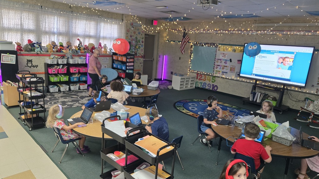 A classroom with children seated at tables, using laptops. A teacher stands behind them, and a projector screen is on the wall.