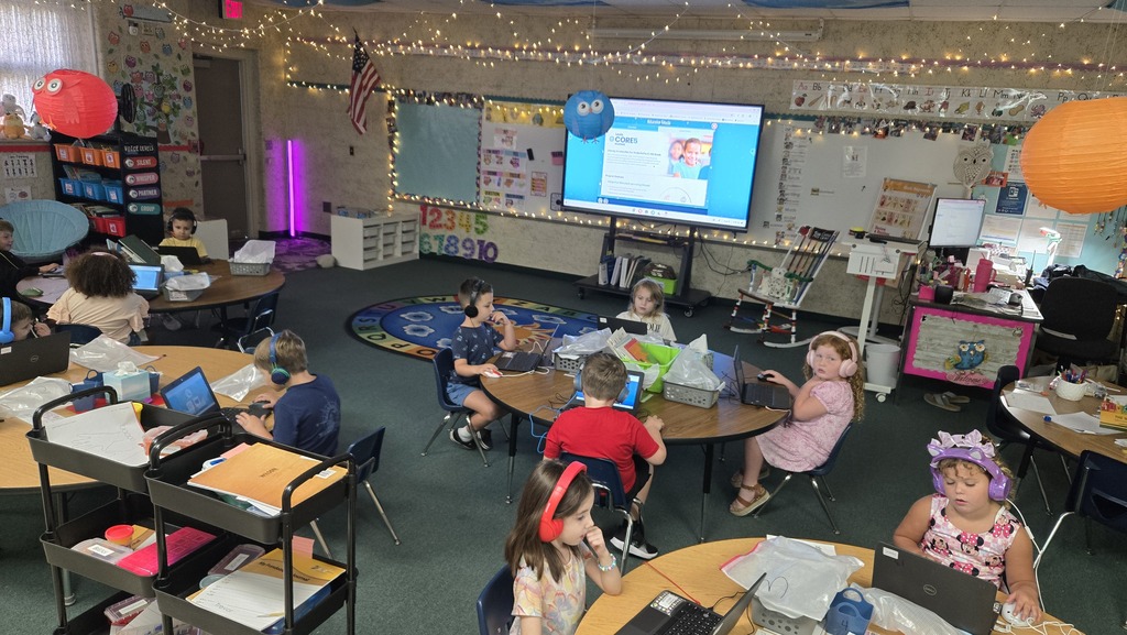 Classroom with children seated at tables, each using laptops. A large monitor is mounted on the wall.