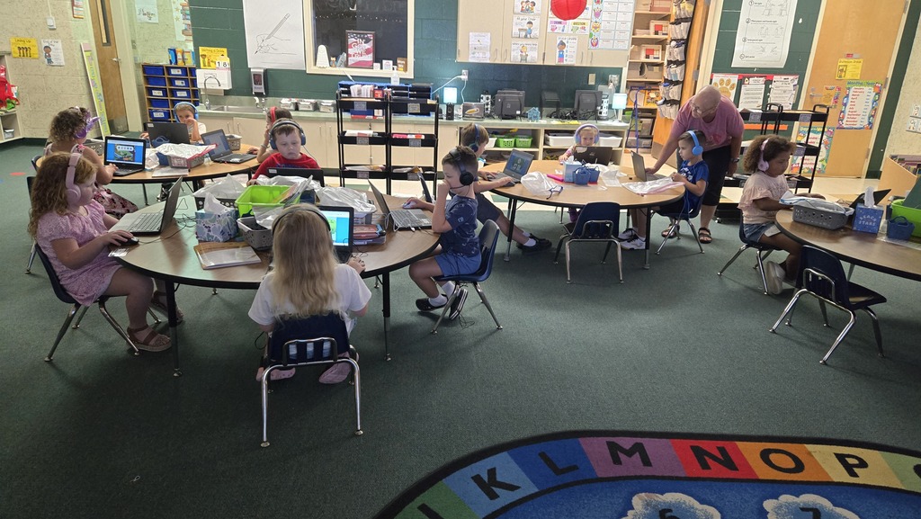 A classroom with children sitting at tables using laptops. Tables are round, chairs are plastic, and a floor mat with letters is visible.