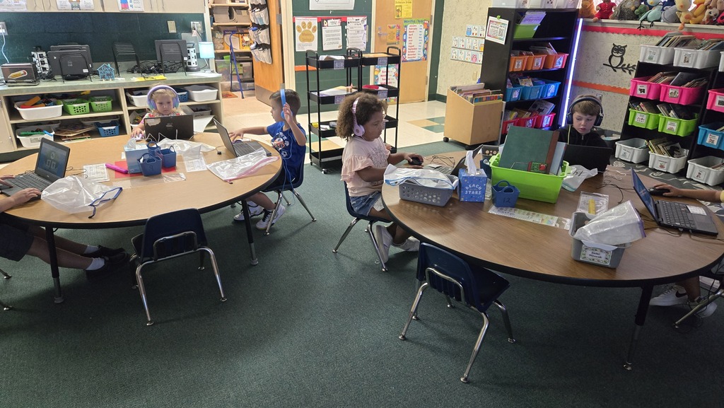 Small children work on laptops at tables in a classroom. Colorful storage bins are behind them.