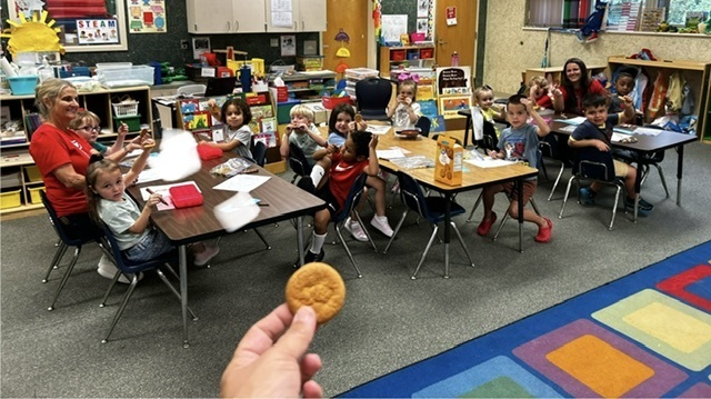Classroom with children seated at desks, a teacher in red shirt holds a cookie, a colorful rug covers the floor.