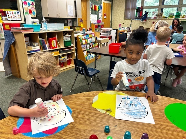Children sit at a table with craft materials. One holds a glue bottle. Shelves and chairs in the background.