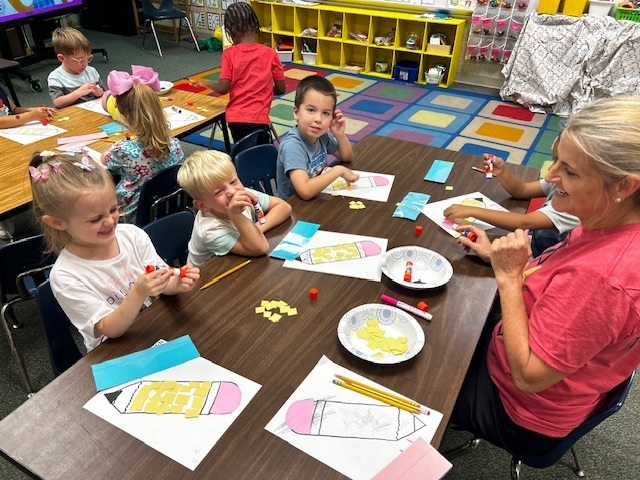 Classroom with children sitting at tables, a woman in front of them, and paper crafts on the table.