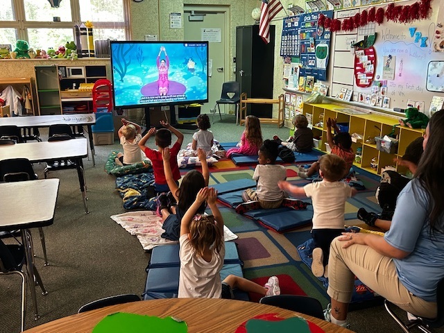 Children sit on colorful mats in a classroom, watching a screen in the foreground. Desks, chairs, and a whiteboard are in the background.