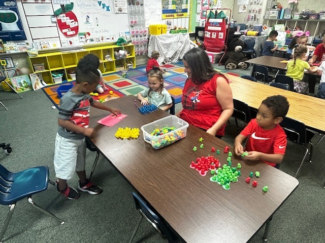 Kids seated at a table with colorful objects, an adult stands next to them. Shelves and books are visible in the background.