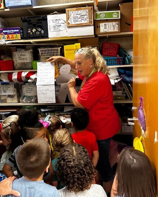 An adult in red stands at a shelf holding paper while children gather around.