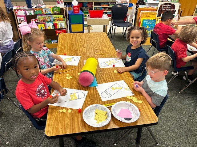 A group of young children sitting at a table, engaging in an activity with paper, scissors, and a pencil.