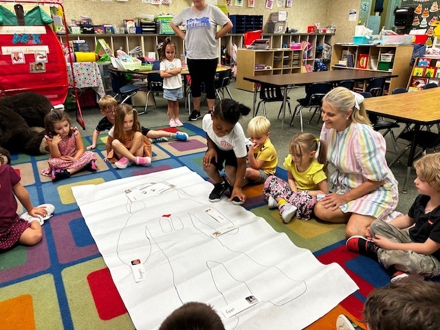 Kids sitting on a multicolored carpet in a classroom with an adult woman. A large diagram is on the floor.