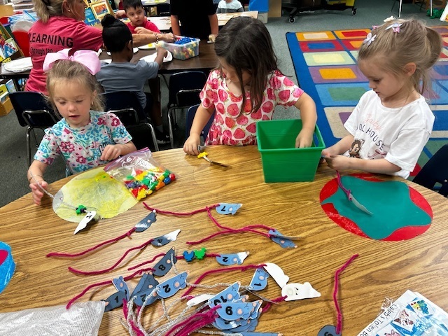 Three young children sit at a round table, engaged in a craft activity with various materials and colorful items.