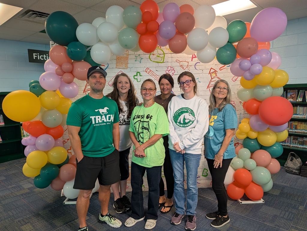 Six individuals in casual clothing pose under a colorful balloon arch in a library.