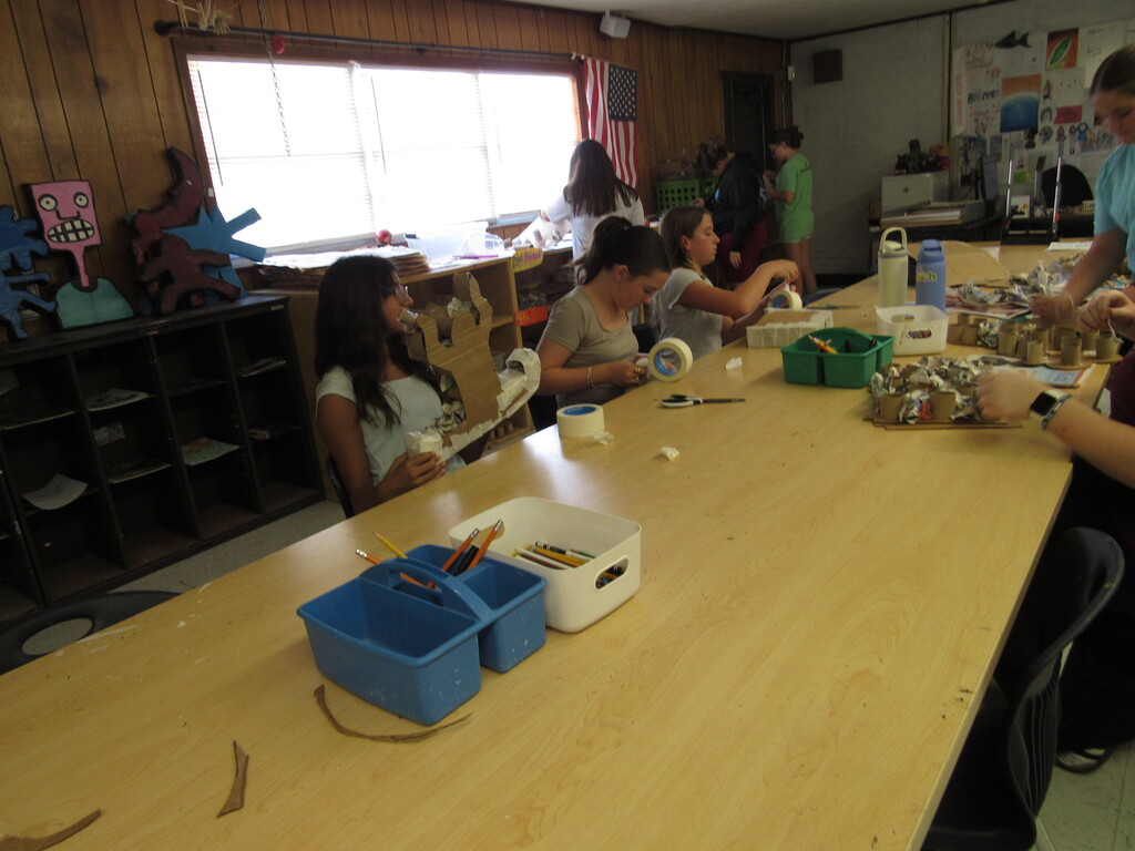 People sit at a table in a classroom with various materials and tools, such as scissors and tape.
