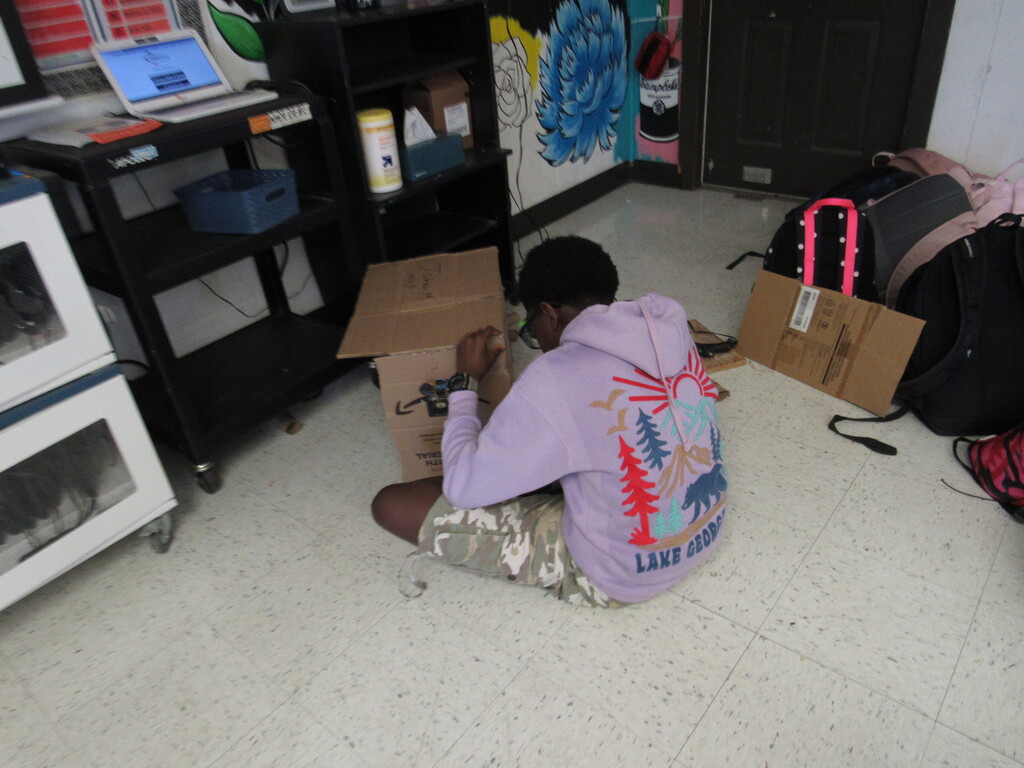 A person in a purple hoodie sits on a floor, working on a box. There are various items in the background.