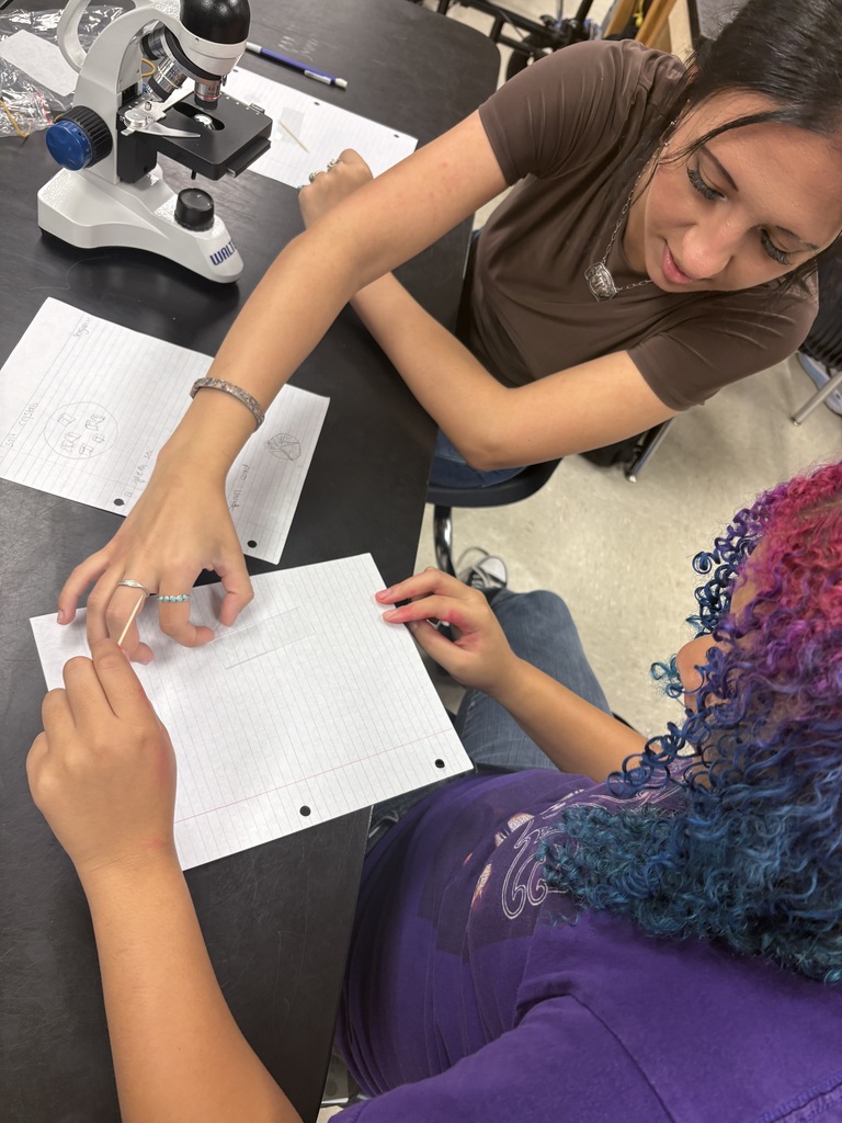 Two individuals are seated, one holding a white paper with text, while the other points at it. A microscope is positioned on the table.