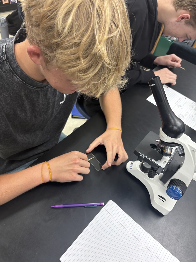 Two students, one focused on a microscope, the other assisting, in a lab setting with papers and a pen on a table.