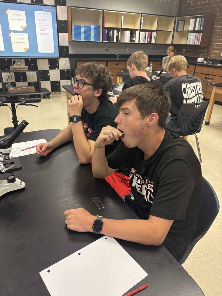 Students sit at a table in a classroom, one using a microscope. Two wear black t-shirts, and one has glasses.
