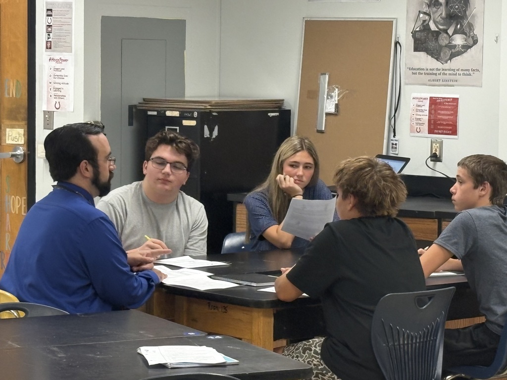 A group of people sit around a table, engaged in discussion. Papers and a laptop are on the table.