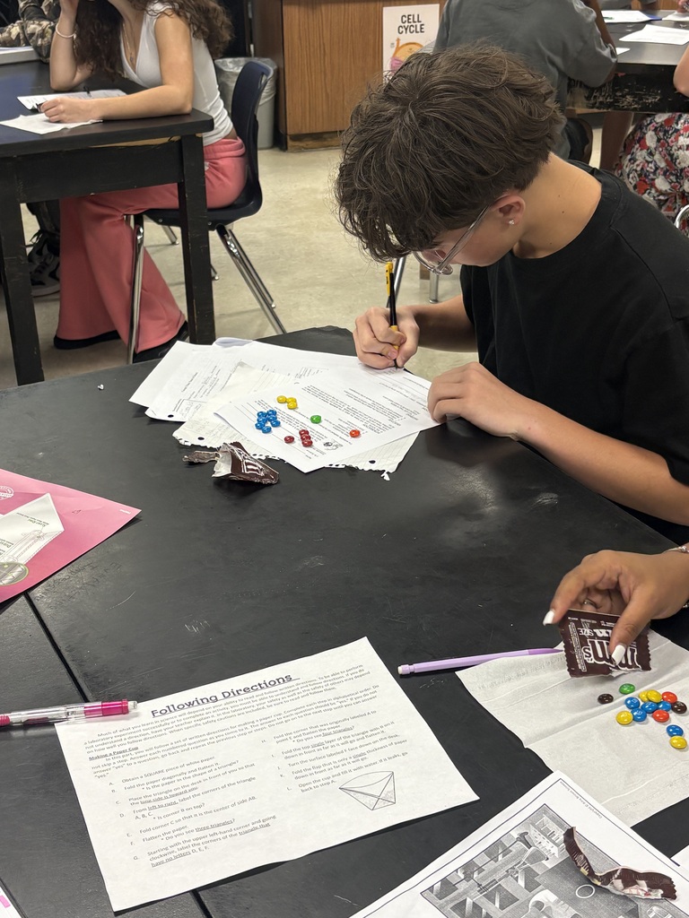 A student with glasses works on a project, using colorful items, a pen, and papers on a black table.