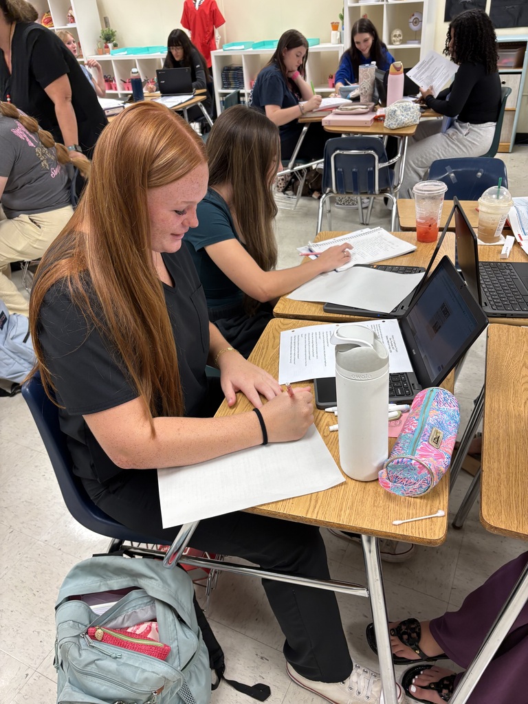 A woman with long hair is seated at a desk, writing on paper. Other students sit nearby.