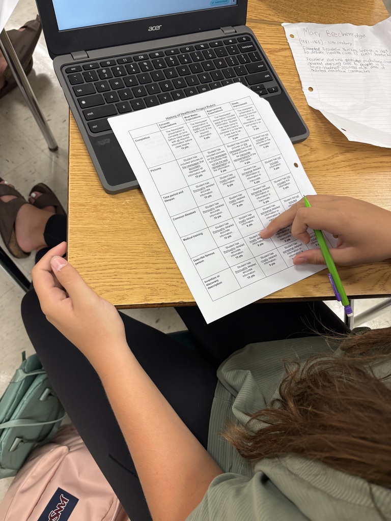 Person seated, holding a worksheet with a pen, a laptop, and other papers on a table.