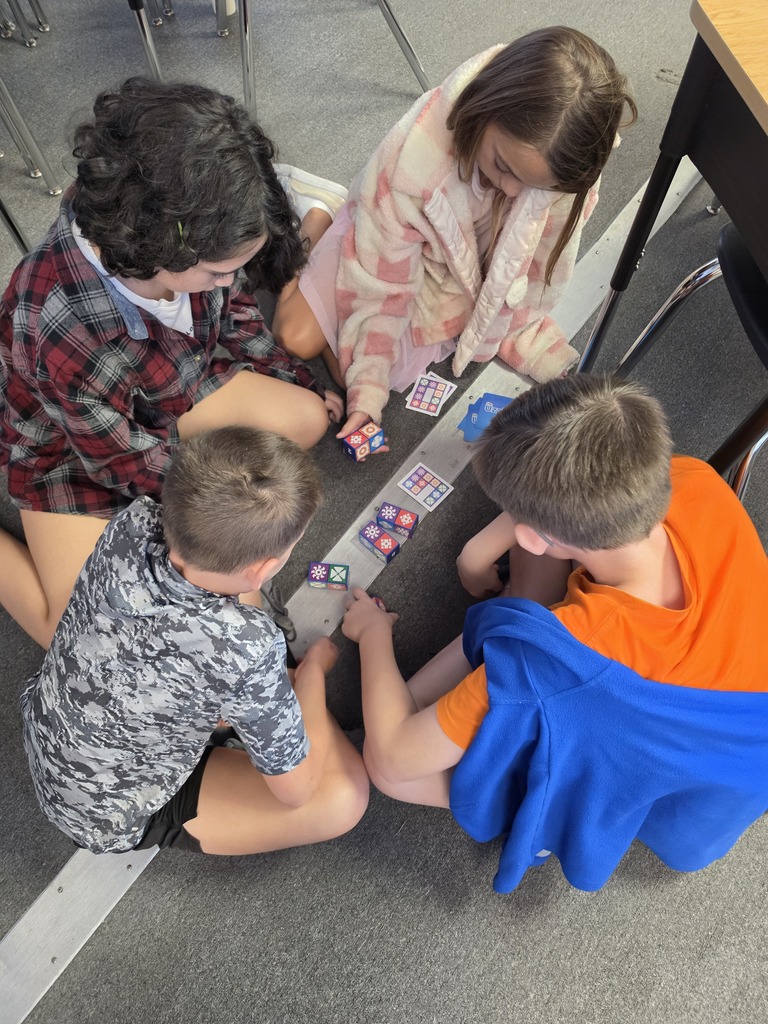Four children sitting on the floor, playing a card game. Two wear shirts, two wear shorts.