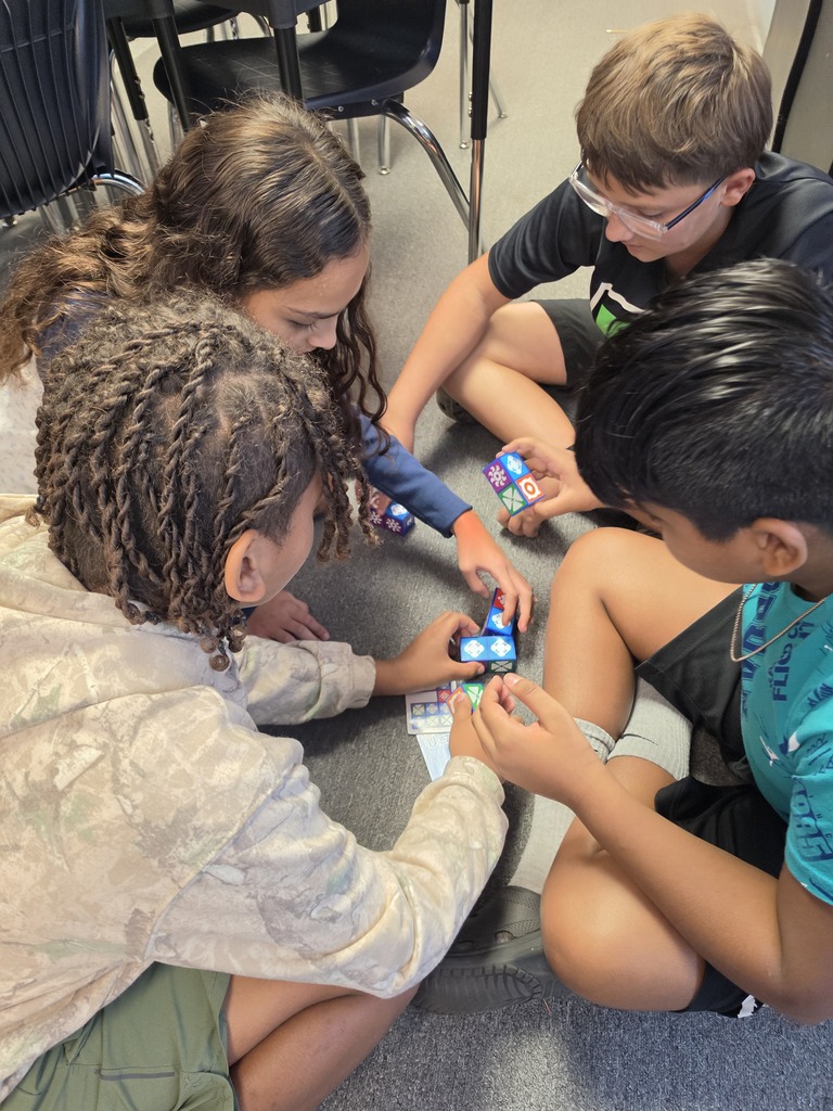 Four children sit on the floor, engaged in a game with colorful cubes. They are in a classroom setting.