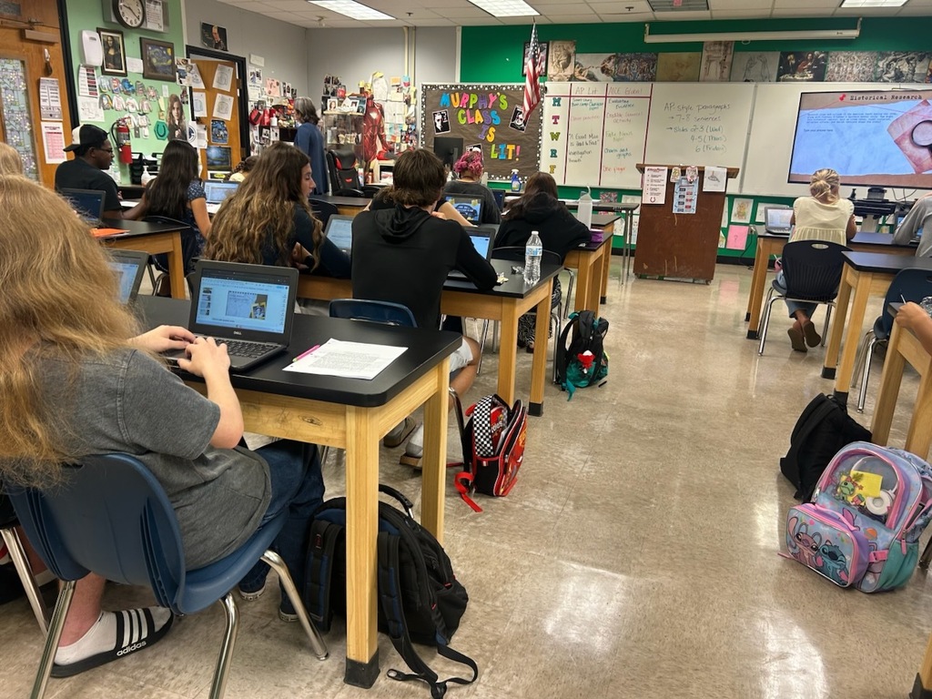 A classroom with students seated at desks, using laptops. A projector screen is visible at the front.