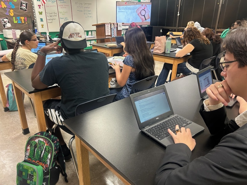 A classroom with students seated at desks, working on laptops. A teacher stands in front of a screen.