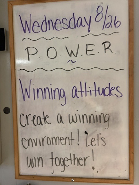 A whiteboard with a brown frame shows motivational text: "Wednesday 8/26, POWER, Winning attitudes create a winning environment! Let's win together!"