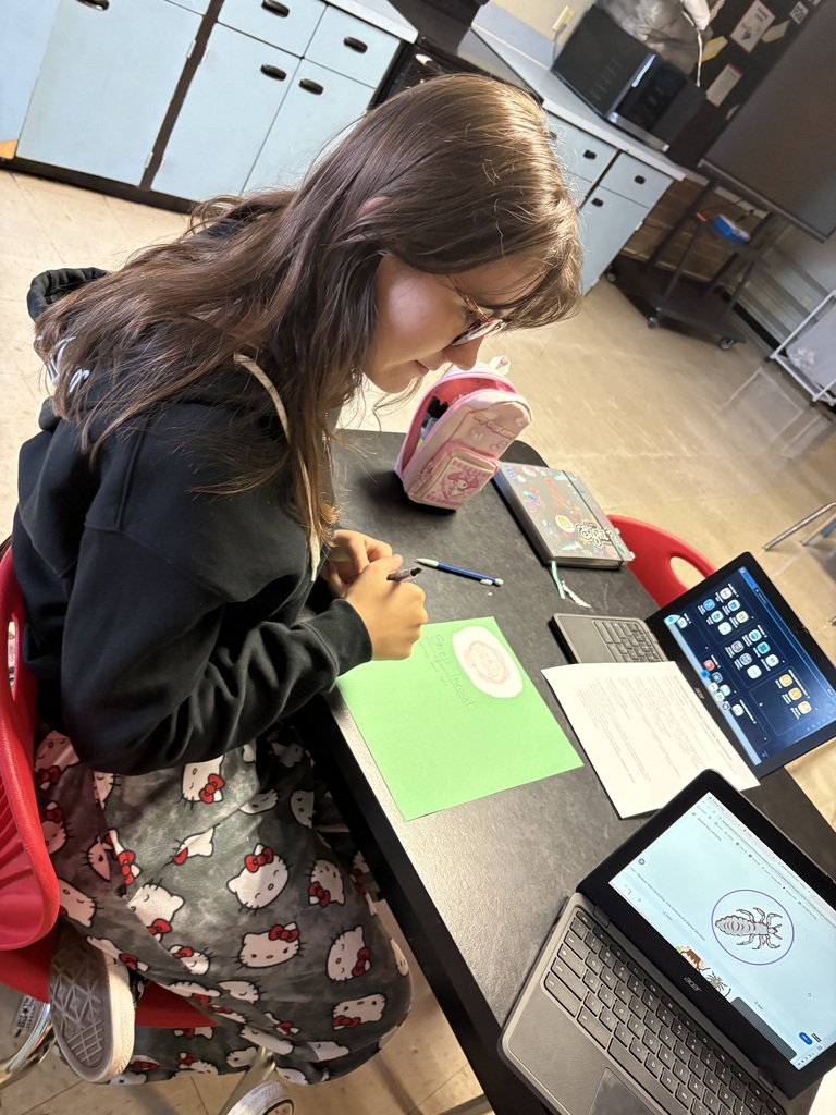 Person at a table working on a green sheet of paper, two laptops open, one with a bee graphic.